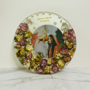 A 10x10 inch circular resin photo frame displaying a preserved wedding photo of a couple, surrounded by real dried white and maroon rose petals, white pearls, and gold text with the names.
